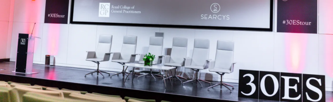 Internal image of meeting space at 30 Euston Square, theatre layout with 5 white chairs set out on a stage, with a lit, branded backdrop, and a lectern.