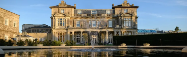 Front, external view of Down Hall Hotel on a sunny day, with clear blue sky, and a fountain in front, reflecting the view of the hotel in its water.