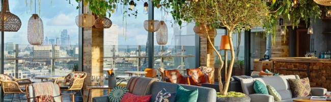 Internal view of relaxed looking space inside Treehouse London - it's a room with colourful and comfy looking sofas and rattan chairs, drop lights, lots of greenery, and big windows with views of the London skyline.